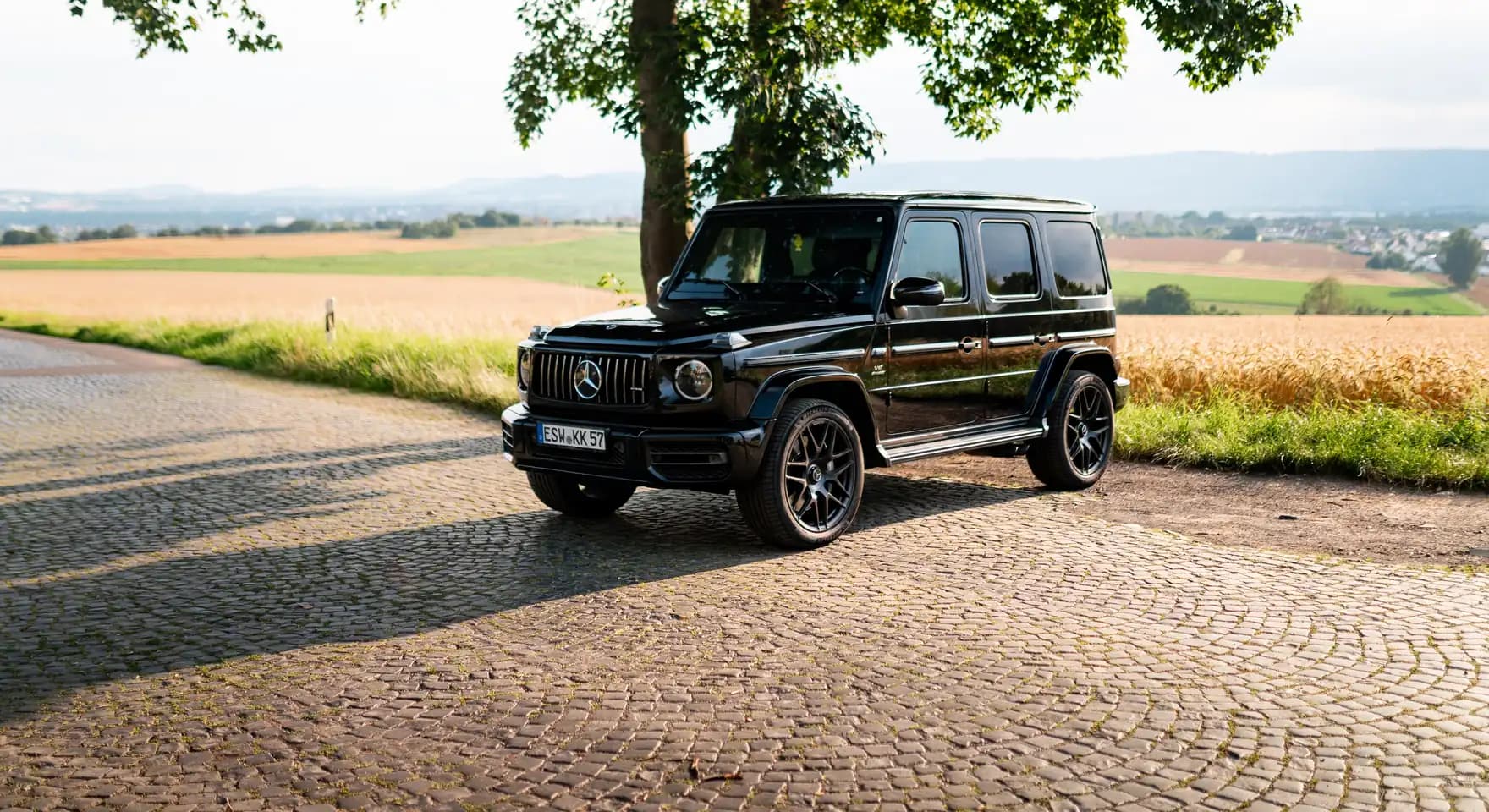 Schwarze Mercedes-AMG G-Klasse unter einem Baum an einem gepflasterten Feldweg, dahinter Felder und Hügel