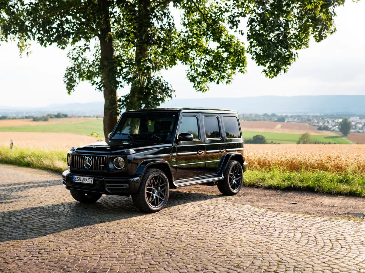 Schwarze Mercedes-AMG G-Klasse unter einem Baum an einem gepflasterten Feldweg, dahinter Felder und Hügel